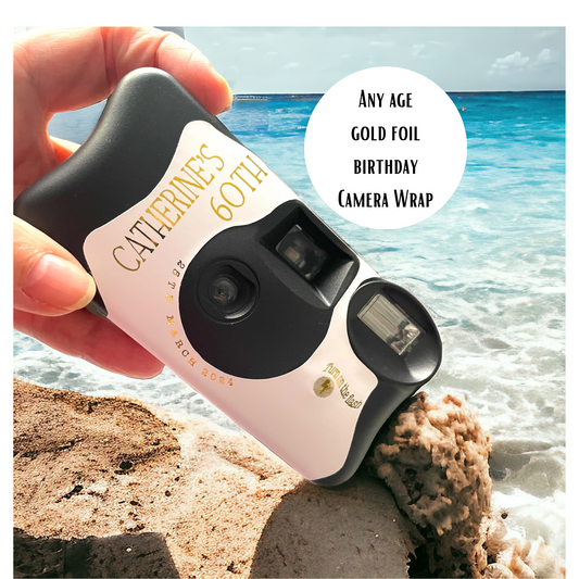 A Kodak FunSaver disposable camera with a replacement personalised sticker for a 60th birthday in gold foil.  Shown on a beach with the sea in the background and a rock and the camera in the foreground.