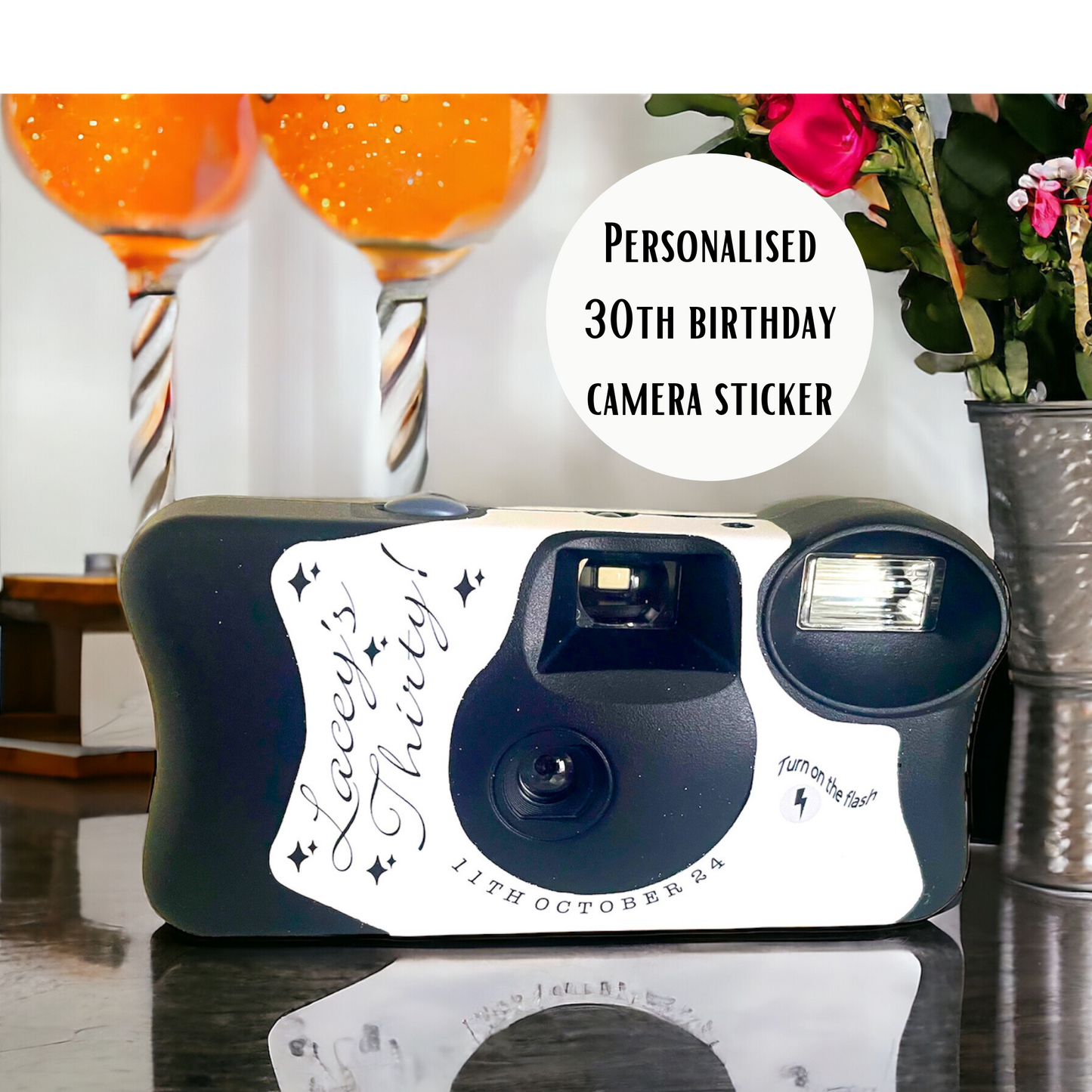 A personalised 30th birthday replacement Kodak FunSaver camera sticker.  Shown pictured on a birthday table with a vase of pink flowers and 2 glass of bubbles,  showing a reflection of the camera in the shiny table.